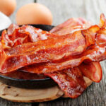 Crispy bacon strips in a cast iron skillet with eggs in the background