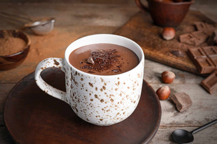 A cup of hot chocolate topped with chocolate shavings and surrounded by hazelnuts