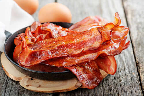 shutterstock_1046058229.jpg Crispy bacon strips in a cast iron skillet with eggs in the background