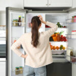 A woman standing in front of an open refrigerator filled with various fruits and vegetables
