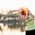 Shocking Back Pain Cure: One Simple Move A woman stretching her arms by a lake during sunrise