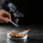 A hand holding a cigarette above an ashtray filled with used cigarettes and smoke rising