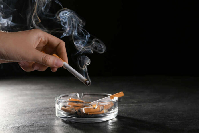A hand holding a cigarette above an ashtray filled with used cigarettes and smoke rising