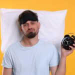 Optimal Sleep Time for Blood Sugar Control Man wearing a sleep mask holding an alarm clock with a frustrated expression