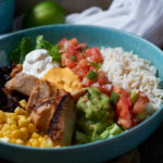 A vibrant bowl filled with grilled chicken, rice, black beans, corn, guacamole, and fresh vegetables