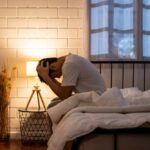 Male Mental Health Crisis: What’s Behind the Mask? A person sitting on a bed with their head in their hands, expressing distress