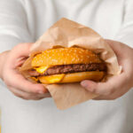 Ultra-Processed Foods: Muscles Filling With Fat? A person holding a cheeseburger wrapped in paper against a yellow background