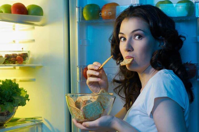 A,Hungry,Girl,Opens,The,Fridge A woman eating a snack while looking at the camera, standing in front of an open refrigerator