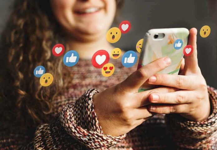 shutterstock_1338120191 Person using smartphone with social media reaction icons floating around