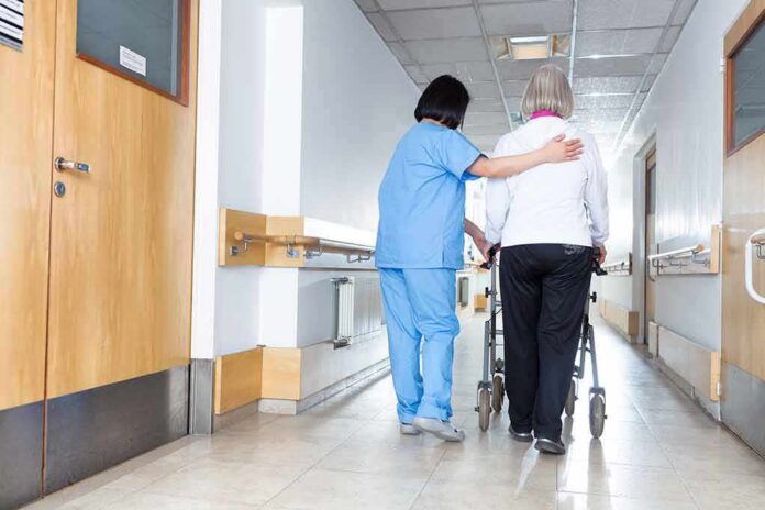 shutterstock_536383975 Nurse assisting an elderly person with a walker in a hallway