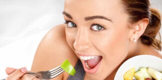 A woman smiling while holding a fork with a piece of green fruit, next to a bowl of mixed fruit.