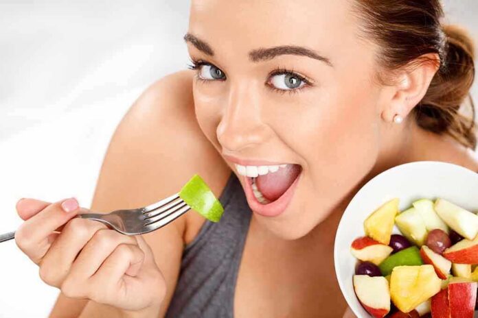 A woman smiling while holding a fork with a piece of green fruit, next to a bowl of mixed fruit.
